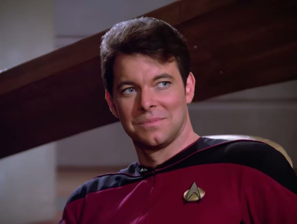 El actor Jonathan Frakes interpreta a William T. Riker en un episodio icónico de la aclamada serie de ciencia ficción Star Trek: La Nueva Generación, con una expresión sonriente. (IMDb)