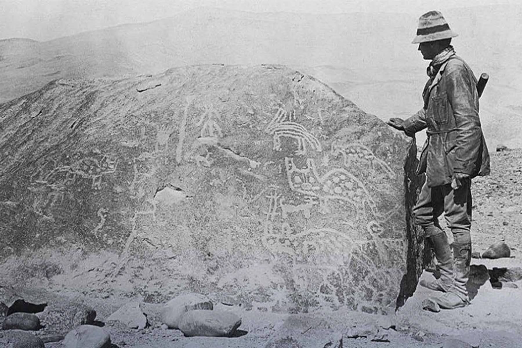 Hiram Bingham III, conocido por sus expediciones en Sudam&eacute;rica, observa una gran roca cubierta de petroglifos que representan figuras zoomorfas y antropomorfas, testimonio del arte rupestre ancestral.