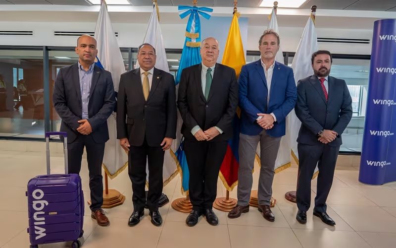 Cinco hombres con atuendo formal se re&uacute;nen en un espacio interior, con banderas nacionales de fondo y un cartel de la aerol&iacute;nea Wingo junto a una maleta morada. (Ministerio de Comunicaciones, Infraestructura y Vivienda)