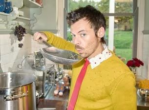 El artista Harry Styles, en una cocina, sostiene una cuchara de servir sobre una olla con un suéter amarillo y una corbata roja. (Harry Styles/RS)