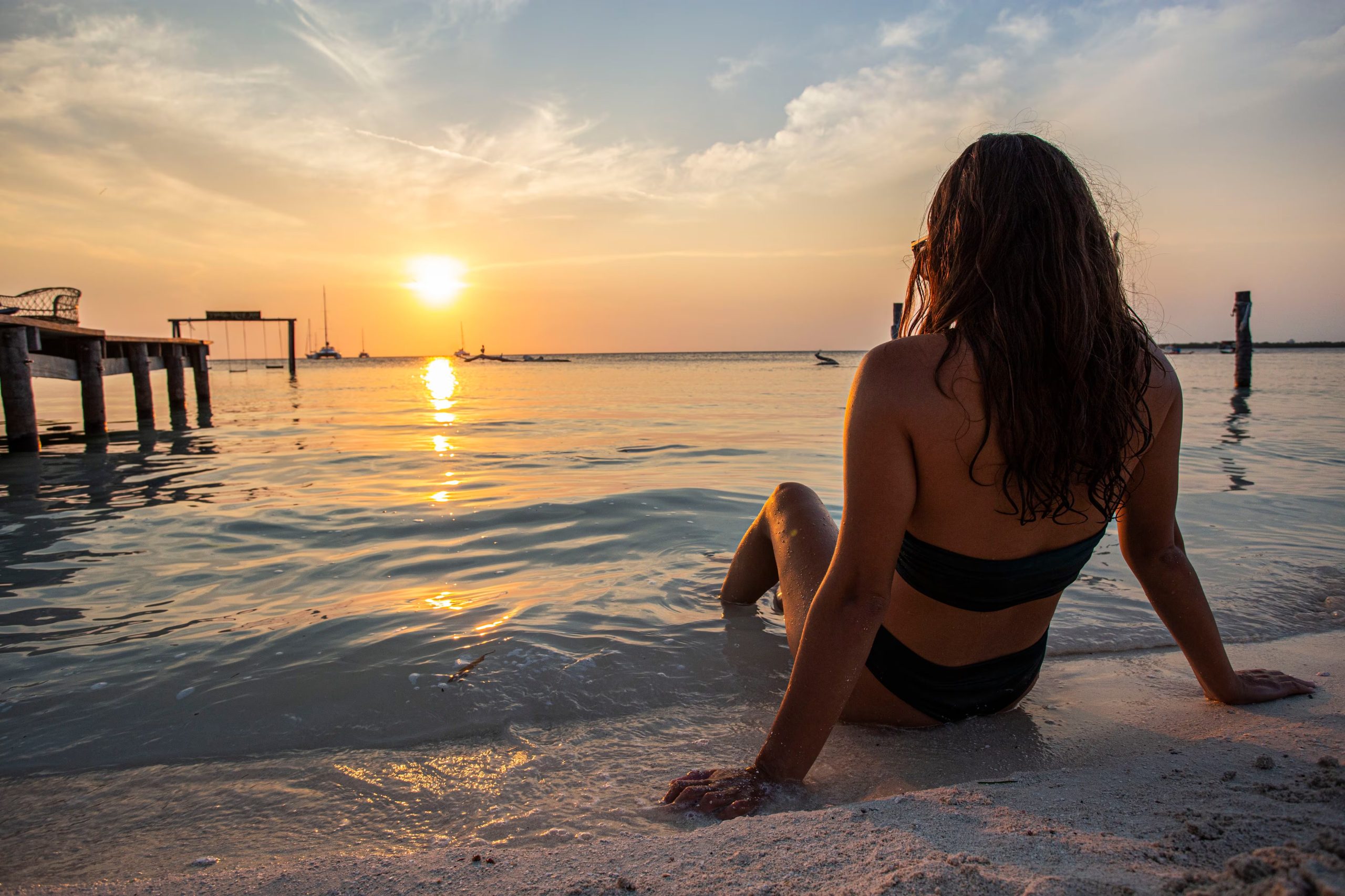 Una mujer de espaldas se sienta en la arena de la playa con los pies en el agua, mirando el sol que se oculta sobre el mar al atardecer. (The Belize Tourism Board (BTB), &oacute;rgano del Ministerio de Turismo y Relaciones con la Di&aacute;spora de Belice.)