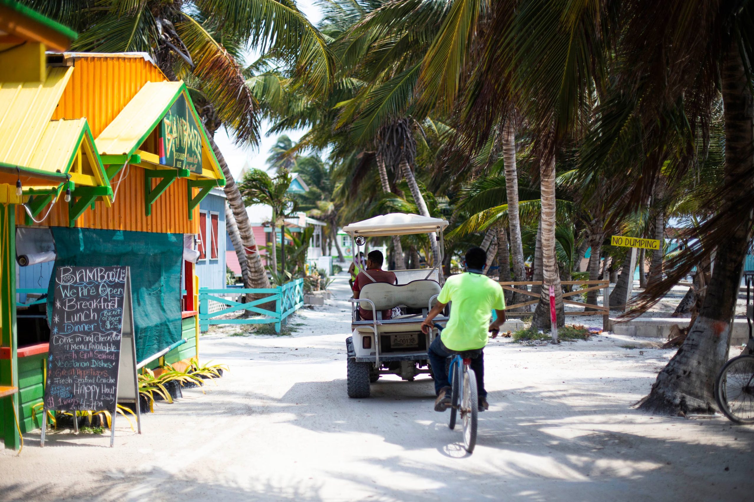 Una calle de arena en un entorno tropical con palmeras muestra a personas que se movilizan en bicicleta y en un carro de golf junto a edificaciones coloridas. (The Belize Tourism Board (BTB), &oacute;rgano del Ministerio de Turismo y Relaciones con la Di&aacute;spora de Belice.)
