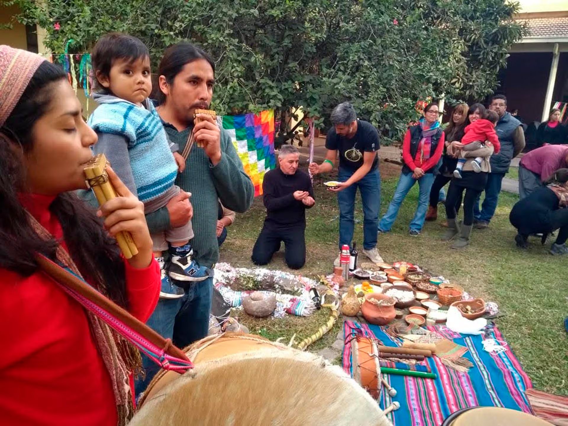 Participantes en la celebración del Día de la Pachamama realizan un ritual con música andina, ofrendas y alimentos en un espacio exterior.